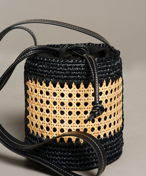 The Tropez Bucket Bag features a cylindrical woven design with black crochet details, a tan rattan center panel, black leather straps, cane handcrafted accents, and a drawstring closure, all set against a plain gray background.