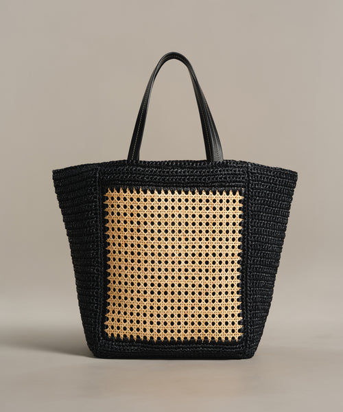The Tropez Tote Bag is a handcrafted summer accessory from Tuscany, highlighting black woven material, dual handles, and a large light cane webbing panel on the front, showcased against a neutral backdrop.