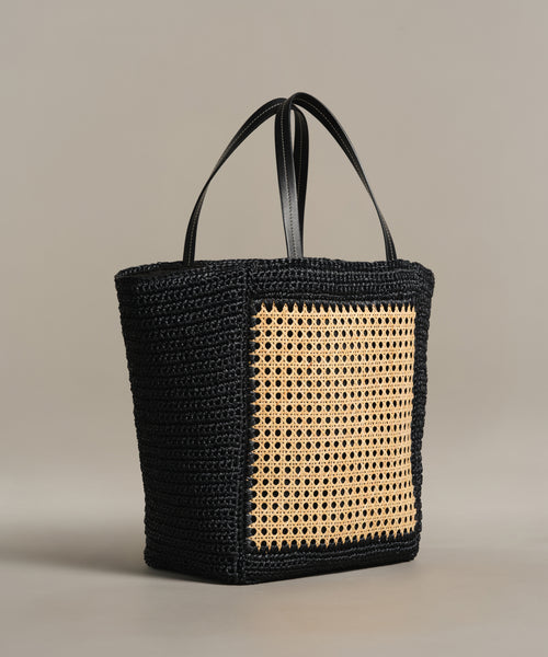 The Tropez Tote Bag, handcrafted in Tuscany, features black woven material, two matching handles, and a prominent tan rattan panel on the front. Its displayed against a plain light grey background.
