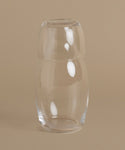 A clear glass bottle with a rounded, tapered shape and a matching glass stopper, set against a solid beige background.