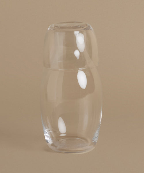 A clear glass bottle with a rounded, tapered shape and a matching glass stopper, set against a solid beige background.