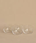 Four clear, round hand-blown bowls from the Universal Low Glass Set of 4 sit in two rows on a beige surface and background. The empty potassium glass bowls feature smooth, minimalist designs inspired by Vincent Van Duysen.