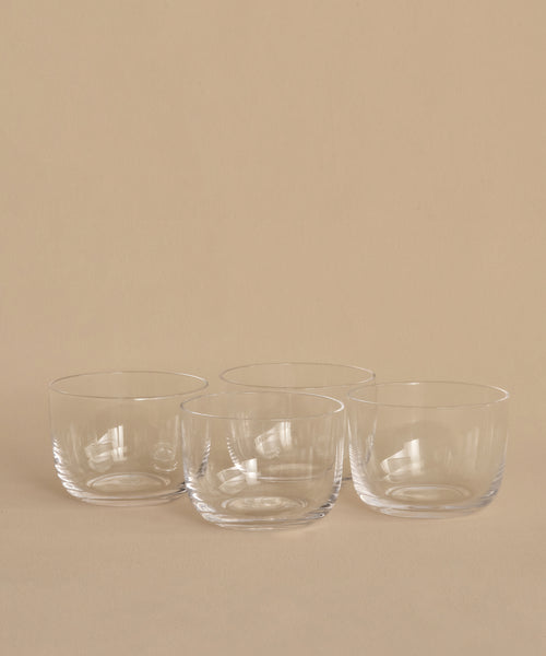 Four clear, round hand-blown bowls from the Universal Low Glass Set of 4 sit in two rows on a beige surface and background. The empty potassium glass bowls feature smooth, minimalist designs inspired by Vincent Van Duysen.