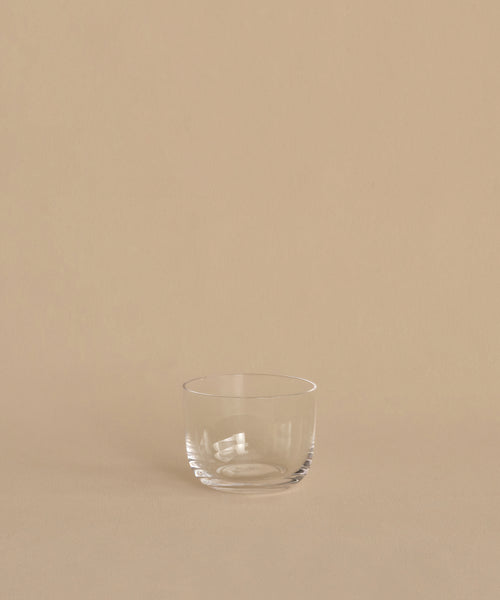 A clear, hand-blown tumbler from the Universal Low Glass Set of 4 rests on a beige surface with a matching background, highlighting Vincent Van Duysen’s minimalist style.