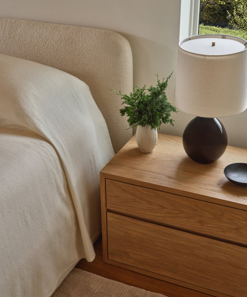 A wooden nightstand sits next to the Upholstered Cove Bed with a cream blanket, topped with a round black-base lamp, small white vase of greenery, and black dish by a window overlooking lush greenery.