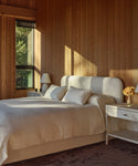 A cozy bedroom with wood-paneled walls and sunlight streaming through a large window features an Upholstered Cove Bed with white bedding. Beside it, tables hold a lamp and a vase of yellow flowers.