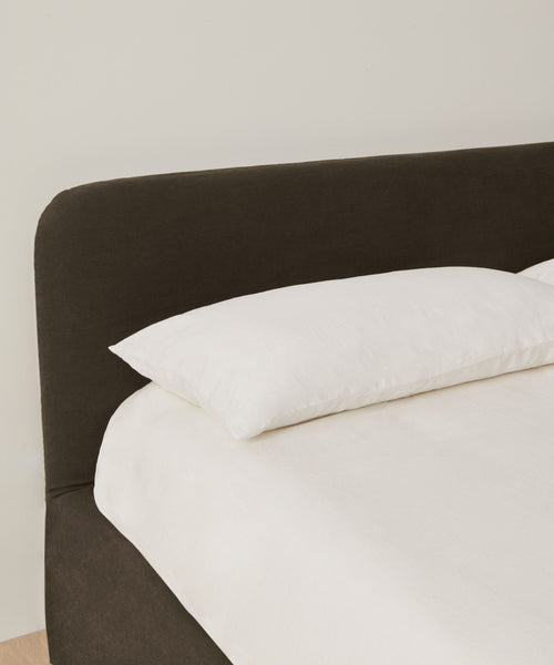 A neatly made bed features a dark upholstered headboard, white pillow, and Jet Linen Cove Bed Slipcover set against a plain light-colored wall.