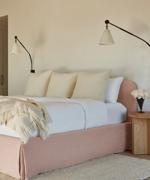 The Cove Bed, featuring a linen-upholstered headboard, is complemented by plush pillows and a bedside lamp to create a serene atmosphere.