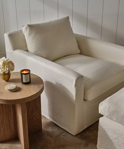 A cozy Harbor Chair in ivory linen with a matching cushion sits by a wooden side table holding a lit candle, white flowers, and a round coaster, set against white paneled walls and beige carpet—classic Jenni Kayne style.