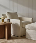 A cozy living space featuring the Harbor Chair in ivory linen with a matching ottoman, a wooden side table holding a lit candle and white flowers, all set against white paneled walls.