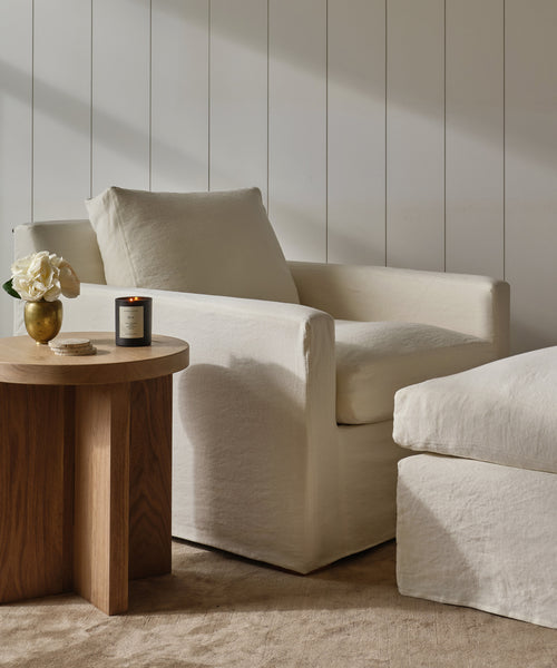 A cozy living space featuring the Harbor Chair in ivory linen with a matching ottoman, a wooden side table holding a lit candle and white flowers, all set against white paneled walls.