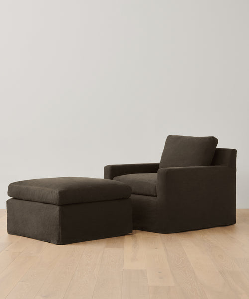 A dark brown Harbor Chair with matching ottoman, both featuring clean lines and plush linen upholstery, are placed on a light wood floor against a plain white wall.