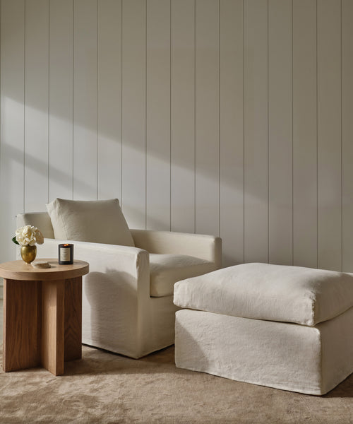 A cozy, modern space featuring the Harbor Ottoman and an Ivory Linen armchair on a beige carpet, next to a round wooden side table with a candle and white flowers, set against a white paneled wall.