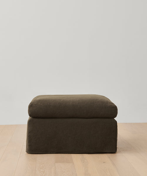 A dark brown Harbor Ottoman with soft, cushioned sides and a rectangular shape sits on a light wood floor in front of a plain white wall.