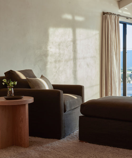 A cozy living room corner with a dark armchair, a Harbor Ottoman from Jenni Kayne Furniture, and a small wooden side table topped with a vase of white flowers. Sunlight streams through light curtains, casting soft shadows on the wall.