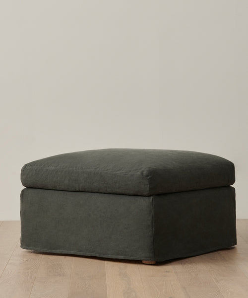 A Harbor Ottoman in dark gray with a plush, cushioned top rests on a light wood floor by an off-white wall, showcasing the refined simplicity of Jenni Kayne Furniture.
