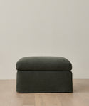 The Harbor Ottoman, upholstered in dark green Slate Linen with a soft cushion, rests on a light wooden floor against a plain, light-colored wall.