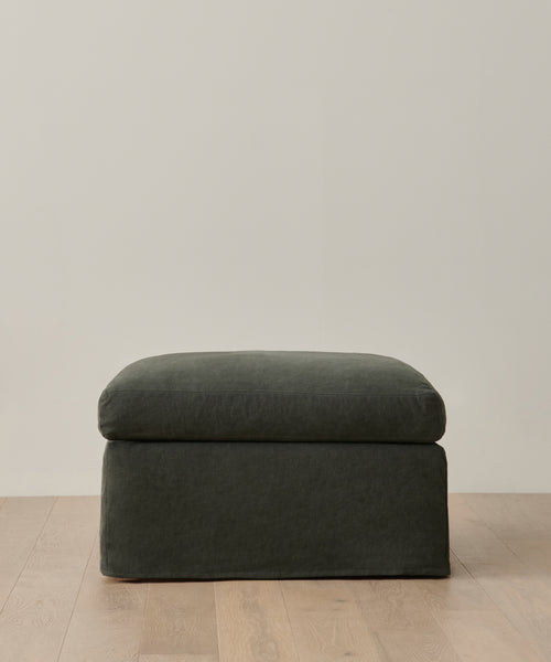The Harbor Ottoman, upholstered in dark green Slate Linen with a soft cushion, rests on a light wooden floor against a plain, light-colored wall.