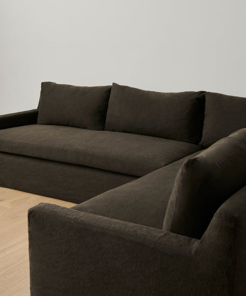 The Harbor Sectional, featuring plush cushions and a modern dark brown design, rests on a light wooden floor against a plain white wall in a minimalist room.