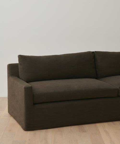A dark brown Harbor Sofa with linen upholstery and clean lines sits on a light wooden floor against a white wall. Only part of this handmade-in-Los Angeles piece, including one armrest and the backrest, is visible.
