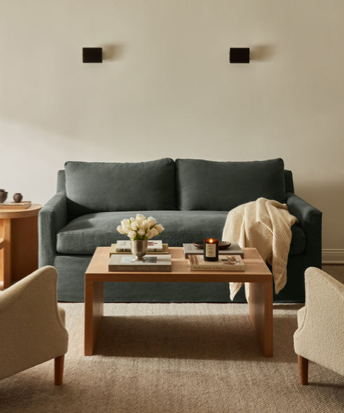 A cozy living room features the dark green linen-upholstered Harbor Sofa, a light throw blanket, a wooden coffee table with books, a candle, and white tulips, two beige armchairs, soft lighting, and cream-colored walls.