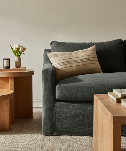 A cozy living room corner highlights the Harbor Sofa with dark gray linen upholstery, paired with a striped beige pillow, light wood coffee tables, white tulips in a vase, a candle, and books on a neutral woven rug against a light wall.