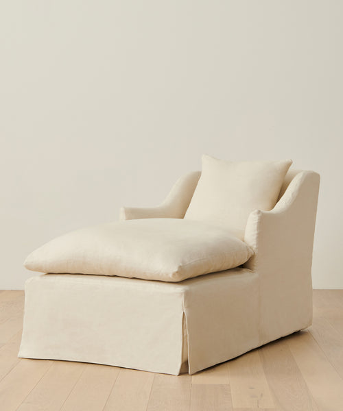 A cream-colored Miramar Chaise with a matching pillow, upholstered in Belgian linen, rests on a light wooden floor against an off-white wall.