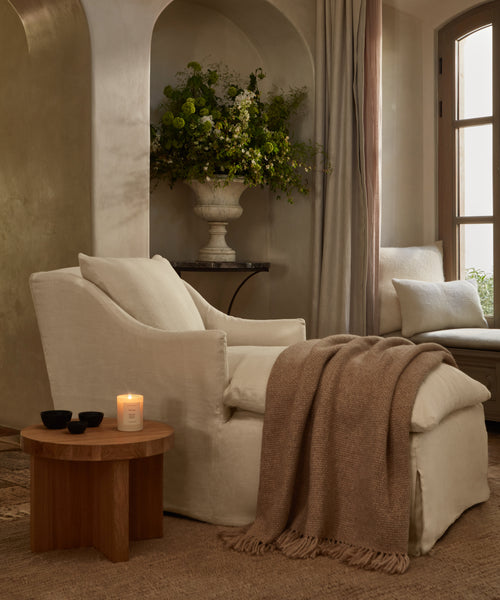 A cream Miramar Chaise with ottoman and brown throw blanket sits by a wooden side table holding a lit candle and black bowls. A large vase of greenery and a window enhance the warm, inviting room.