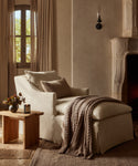 A Miramar Chaise with a throw blanket and pillow is placed by a wooden side table holding a candle and flowers, set in a softly lit room with neutral tones, curtains, and a stone fireplace.