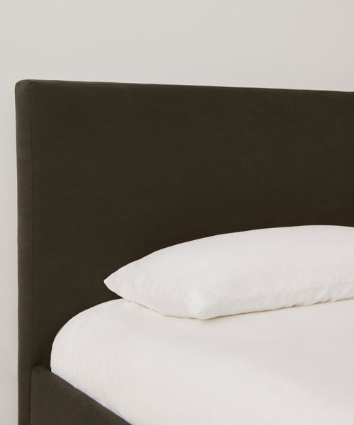 A close-up of the Pacific Bed Warehouse Sale features a dark upholstered headboard, crisp white pillow, and Belgian linen bedding set against a plain light-colored wall.