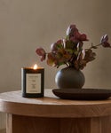 The Vail Glass Candle, a hand-poured black candle, sits alongside a round vase of reddish-purple flowers and an empty dark dish on a round wooden table against a neutral background, creating an inviting holiday scent.