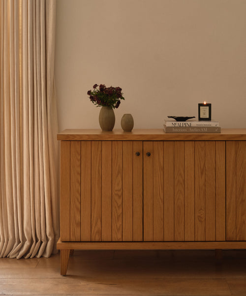 A wooden cabinet with vertical grooves stands against a cream wall. On top are dark flowers in a vase, a small ceramic vessel, books, and the Vail Glass Candle with a holiday scent. Cream curtains hang to the left.