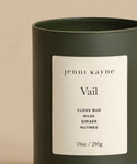 The Vail Glass Candle is a hand-poured dark green glass candle with a white jenni kayne Vail label, featuring clove bud, musk, ginger, and nutmeg scents. 10oz/295g—an inviting holiday aroma set against a beige background.