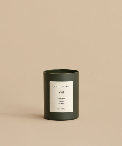 A minimalist Vail Glass Candle, hand-poured with a holiday scent, features a white label reading Jenni Kayne Vail Clove Bud & Siberian Juniper, 10oz / 283g, set against a beige background.