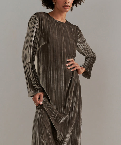A person wearing the Velvet Stella Dress, a long-sleeved, metallic pleated holiday dress in dark, shimmery fabric, poses with one hand on their hip and the other holding the skirt against a plain, neutral background.