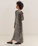 A woman with short curly hair models the Velvet Stella Dress—an ankle-length, long-sleeve holiday dress in silver-grey crushed velvet with vertical pleats—paired with black flats, standing sideways against a plain background.