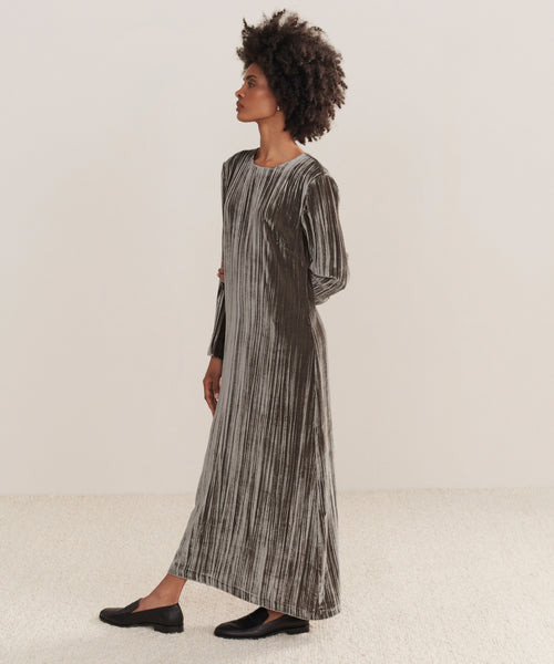 A woman with short curly hair models the Velvet Stella Dress—an ankle-length, long-sleeve holiday dress in silver-grey crushed velvet with vertical pleats—paired with black flats, standing sideways against a plain background.