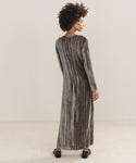 A woman with curly hair faces away, wearing the Velvet Stella Dress—a long, loose-fit silver-gray crushed velvet holiday dress—on a light carpeted floor before an off-white background.