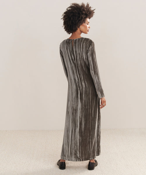 A woman with curly hair faces away, wearing the Velvet Stella Dress—a long, loose-fit silver-gray crushed velvet holiday dress—on a light carpeted floor before an off-white background.