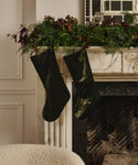 Two Velvet Stockings hang from a marble fireplace mantel decorated with green foliage and red-brown flowers, bringing classic holiday charm to the scene. A mirror and window reflection are visible above.