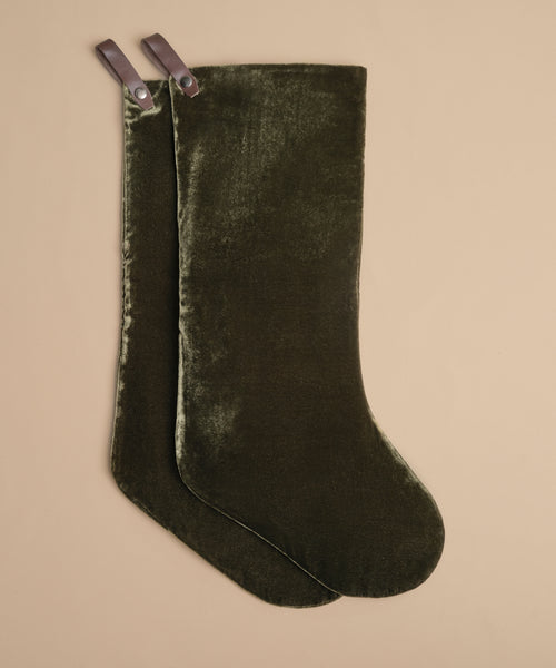 Two Velvet Stockings in dark olive with leather hanging loops lie flat on a beige surface, slightly overlapping—an elegant addition to your holiday décor.