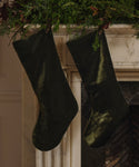 Two Velvet Stockings in dark olive velvet hang from a decorated white marble mantle, surrounded by lush green foliage for classic holiday décor.