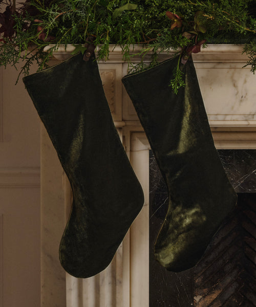 Two Velvet Stockings in dark olive velvet hang from a decorated white marble mantle, surrounded by lush green foliage for classic holiday décor.