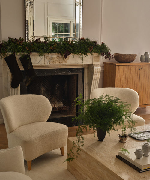 A cozy living room features light armchairs, a marble coffee table, and a fireplace decorated with garland and Velvet Stockings in dark olive. A leafy plant sits on the table, complemented by a mirror and warm wood furniture for festive charm.