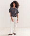 A woman with voluminous curly hair stands against a plain background, wearing the Vintage Pocket Tee in charcoal gray. She pairs it with white pants, a black belt, and black sandals, resting one hand in her pocket as she gazes calmly at the camera.