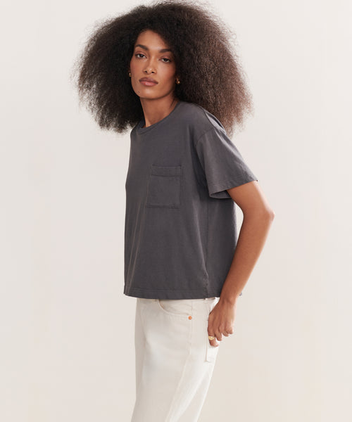Wearing the Vintage Pocket Tee in charcoal, a person with voluminous curly hair pairs it with light jeans, standing against a plain background for an effortless casual look.