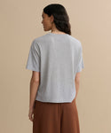 A woman with long, dark hair stands with her back to the camera, wearing a Vintage Pocket Tee in Heather Grey and high-waisted brown pants against a beige background.