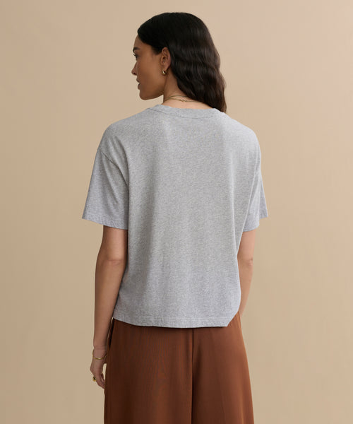 A woman with long, dark hair stands with her back to the camera, wearing a Vintage Pocket Tee in Heather Grey and high-waisted brown pants against a beige background.