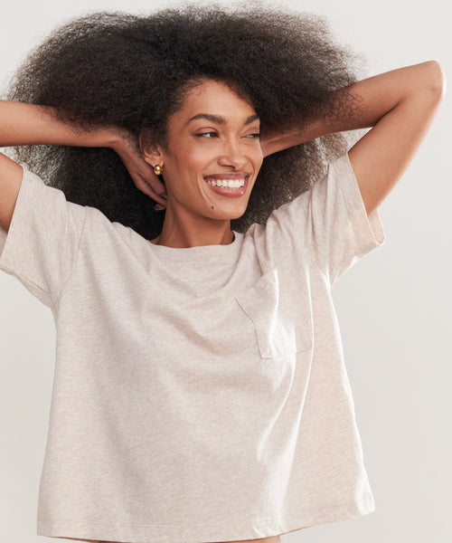 A woman with voluminous curly hair smiles with her arms raised, wearing the Vintage Pocket Tee—a light beige, short-sleeve top with a chest pocket for a casual look.