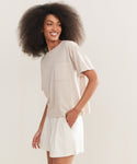 A woman with voluminous curly hair smiles in a Vintage Pocket Tee and white shorts, standing against a plain light background for a relaxed, casual look.